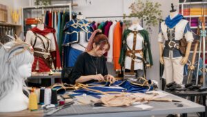 A bright cosplay workshop with costume pieces, colorful fabric, props, wigs, mannequins, and gaming controllers.
